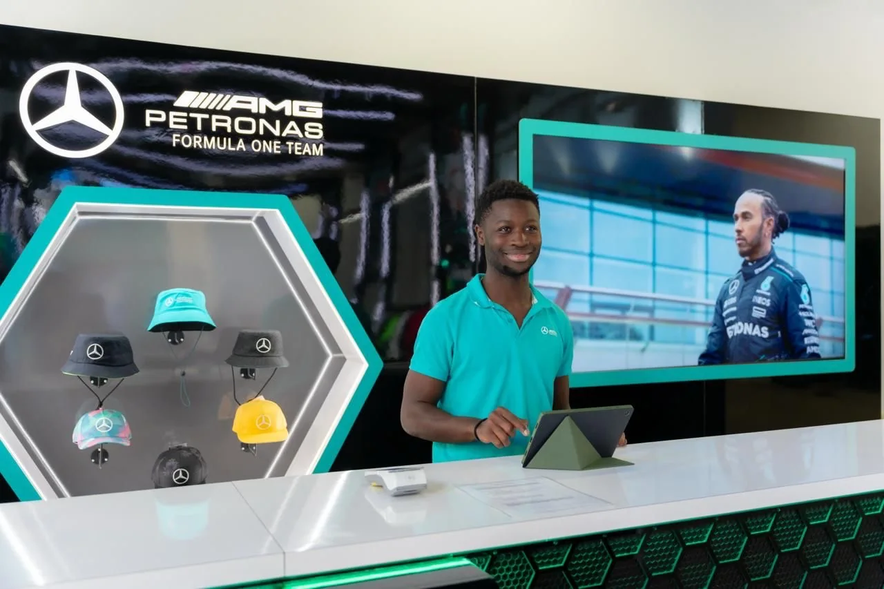 Miembro del equipo de sala en el mostrador, bajo el rótulo de Mercedes-AMG PETRONAS y un expositor hexagonal de gorras